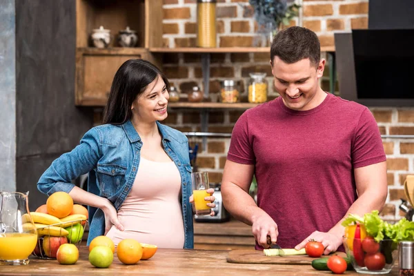 Sonriente joven embarazada sosteniendo un vaso de jugo fresco y mirando al marido cortando apio en la cocina - foto de stock
