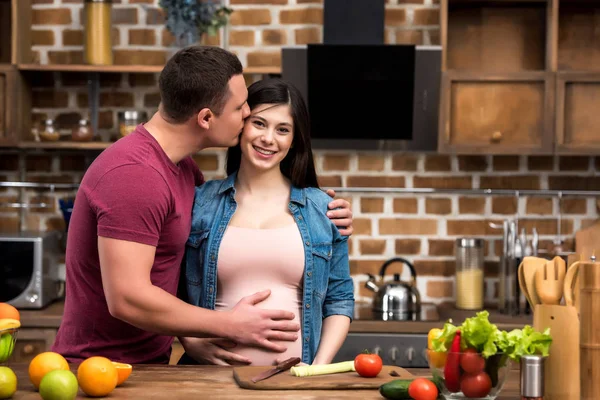 Joven abrazando y besando feliz esposa embarazada en la cocina - foto de stock