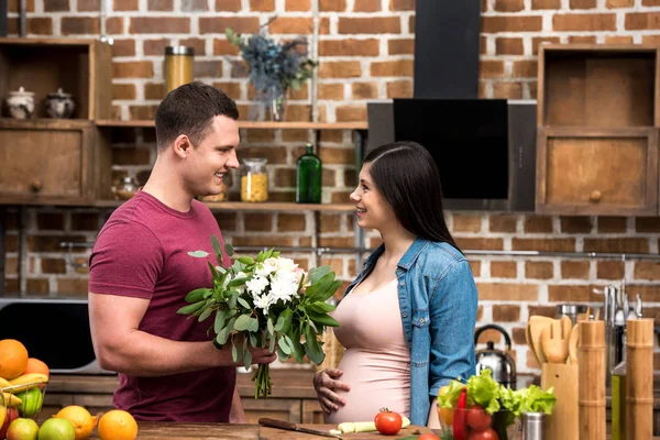 Vue latérale du jeune homme souriant présentant un bouquet de fleurs à une femme enceinte heureuse — Photo de stock