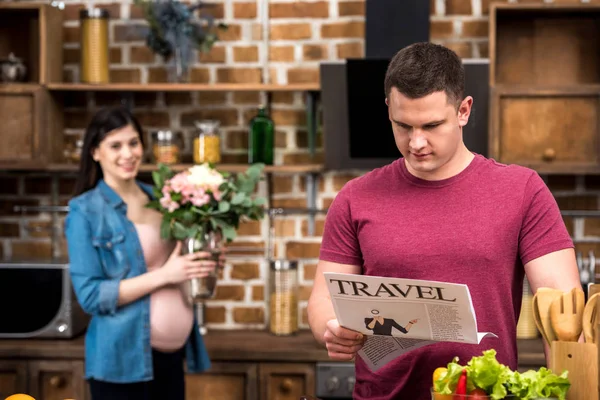 Giovane uomo leggere giornale di viaggio mentre felice moglie incinta in possesso di vaso con fiori dietro — Foto stock