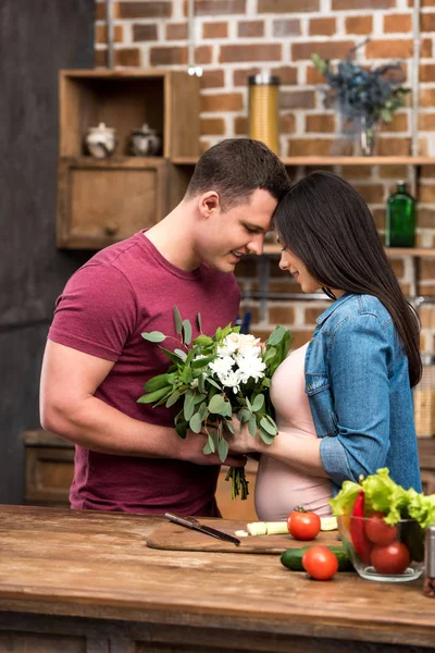 Glücklicher junger Mann und schwangere Frauen berühren Stirn und halten schöne Blumen zu Hause — Stockfoto