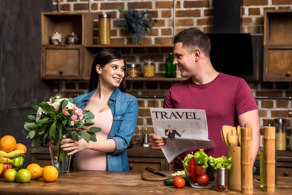 Smiling young man holding travel newspaper and looking at happy pregnant wife arranging flowers in vase — Stock Photo