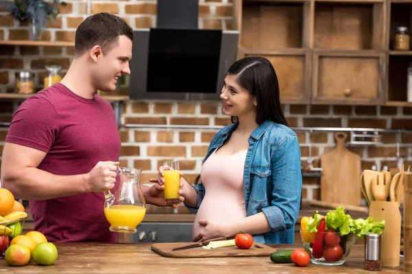 Счастливая молодая беременная пара держит стакан и кувшин свежего апельсинового сока и улыбается друг другу на кухне — стоковое фото