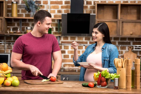 Feliz joven embarazada pareja sonriendo entre sí mientras cocinar juntos - foto de stock