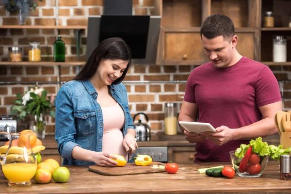 Улыбающаяся молодая беременная женщина режет перец и мужчина с помощью цифрового планшета на кухне — стоковое фото
