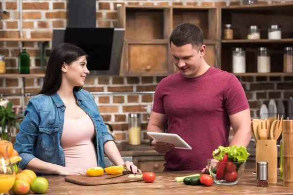 Happy young pregnant couple preparing fresh vegetable salad, man using digital tablet — Stock Photo