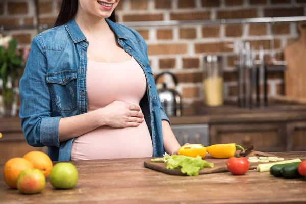 Обрізаний знімок усміхненої молодої вагітної жінки, яка торкається живота під час приготування їжі — стокове фото