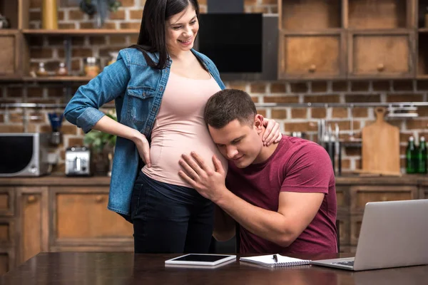 Young man working at home and listening belly of happy pregnant wife — Stock Photo