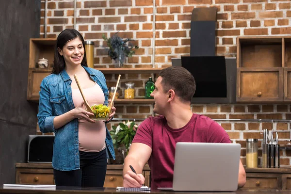 Jovem sorrindo usando laptop e olhando para a esposa grávida segurando tigela de vidro com salada de legumes — Fotografia de Stock