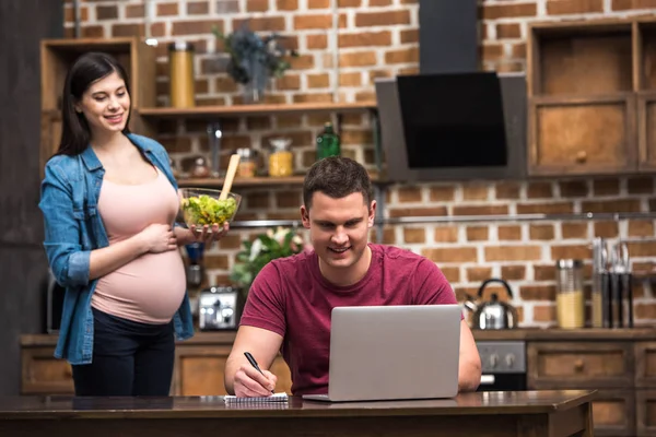 Souriant jeune homme à l'aide d'un ordinateur portable et de prendre des notes tandis que la femme enceinte tenant bol en verre avec salade de légumes — Photo de stock