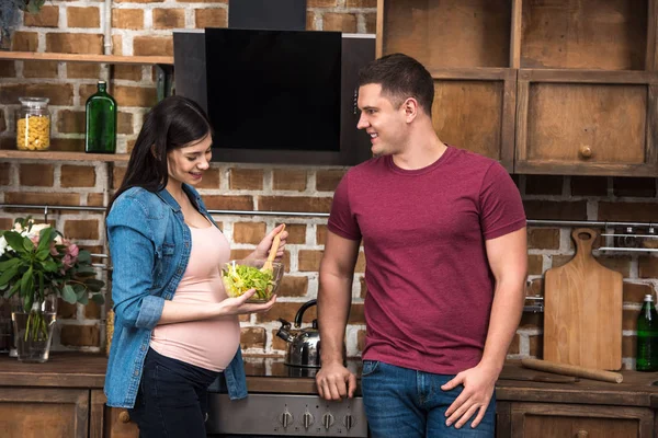 Glücklicher junger Mann blickt lächelnde schwangere Frau an, die Schüssel mit Gemüsesalat in der Küche hält — Stockfoto