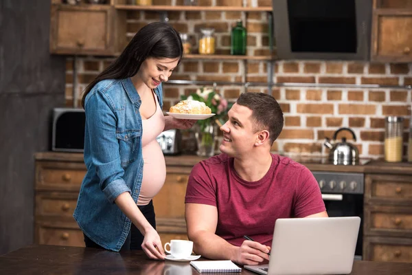 Lächelnde junge schwangere Frau gibt Kaffee und Gebäck an glücklichen Ehemann, der zu Hause mit Laptop arbeitet — Stockfoto