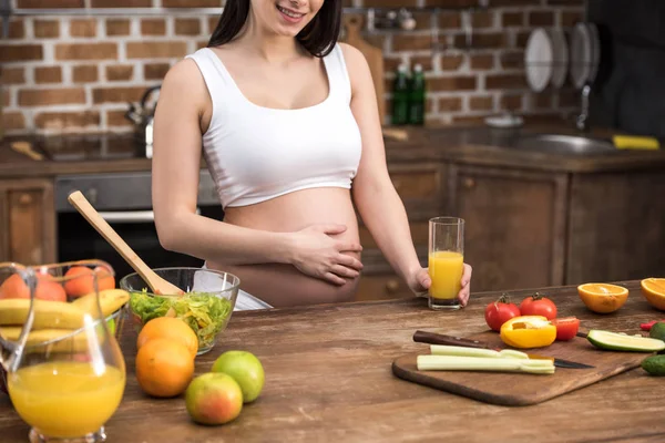 Recortado disparo de sonriente joven embarazada tocando vientre y sosteniendo vaso de jugo fresco en la cocina - foto de stock