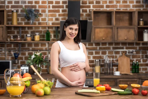 Hermosa joven embarazada tocando el vientre y sonriendo a la cámara en la cocina - foto de stock