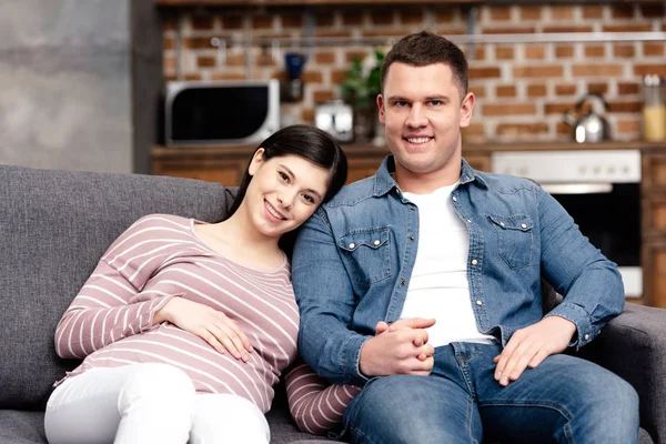Счастливая молодая беременная пара, сидящая вместе на диване и улыбающаяся в камеру — стоковое фото