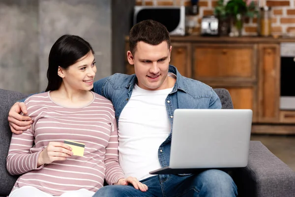 Feliz joven pareja embarazada con tarjeta de crédito utilizando el ordenador portátil juntos en casa - foto de stock