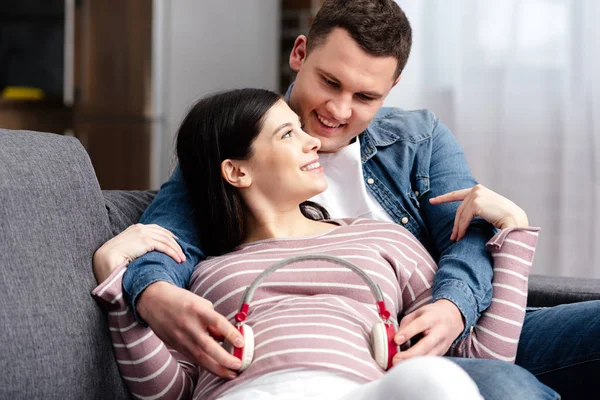 Feliz joven pareja embarazada usando auriculares para el vientre en casa - foto de stock