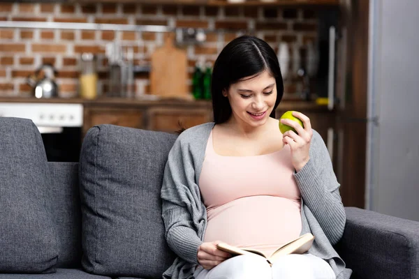 Sonriente joven embarazada comer manzana y leer libro en casa - foto de stock