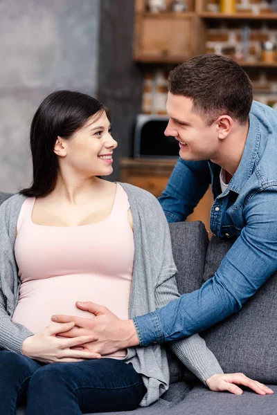 Glückliches junges schwangeres Paar berührt Bauch und lächelt sich zu Hause an — Stockfoto