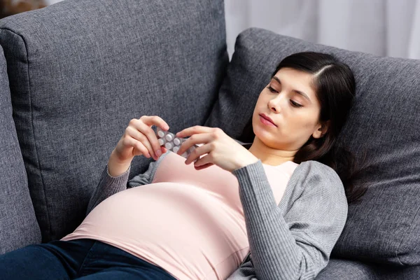 Jovem grávida segurando medicina enquanto deitado no sofá em casa — Fotografia de Stock