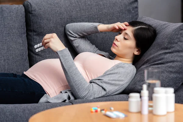 Молода вагітна жінка тримає таблетки, лежачи на дивані вдома — стокове фото