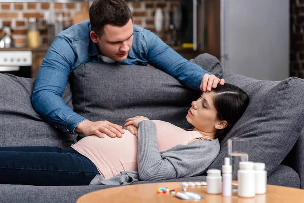 Молодой человек касается лба и живота больной беременной женщины, лежащей на диване — стоковое фото