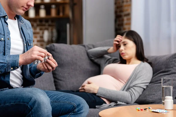 Recortado tiro de hombre sosteniendo píldoras mientras enfermo embarazada acostado en sofá - foto de stock