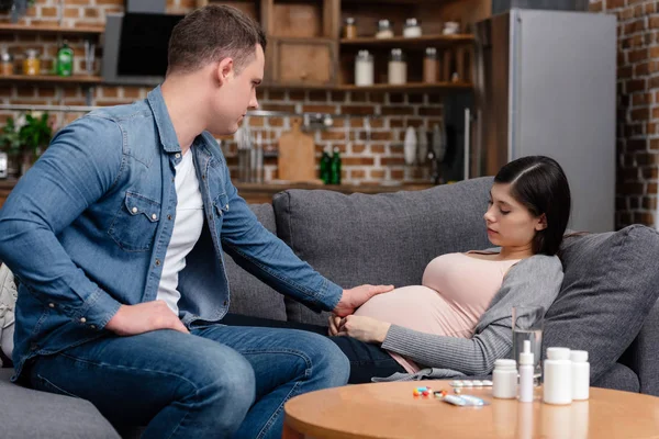 Man touching belly of pregnant woman lying on sofa and various pills on table — Stock Photo