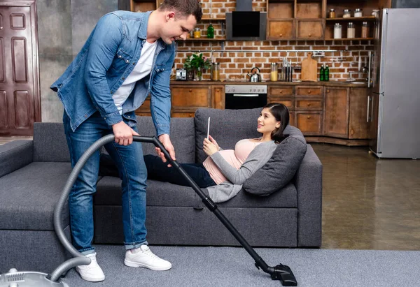 Hombre joven limpieza de alfombras con aspiradora durante el embarazo mujer acostada con la tableta digital en el sofá - foto de stock