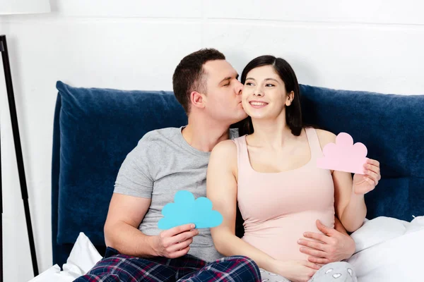 Feliz jovem grávida casal segurando azul e rosa nuvens símbolos e beijos na cama — Fotografia de Stock
