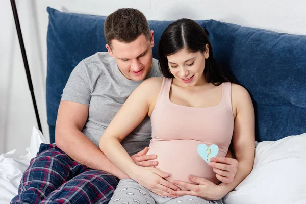 Счастливая молодая беременная пара трогает живот и держит карточку в форме сердца с вопросительным знаком — стоковое фото