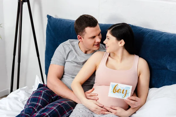 Glückliches junges schwangere Paar lächelt einander auf dem Bett und hält Karte mit Wort Junge — Stockfoto