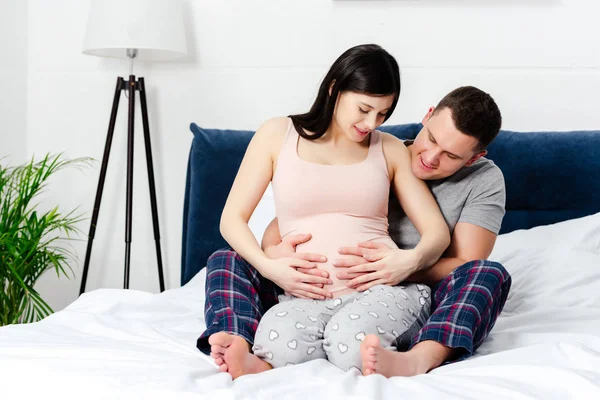 Счастливая молодая беременная пара сидит на кровати и трогает животик вместе — стоковое фото