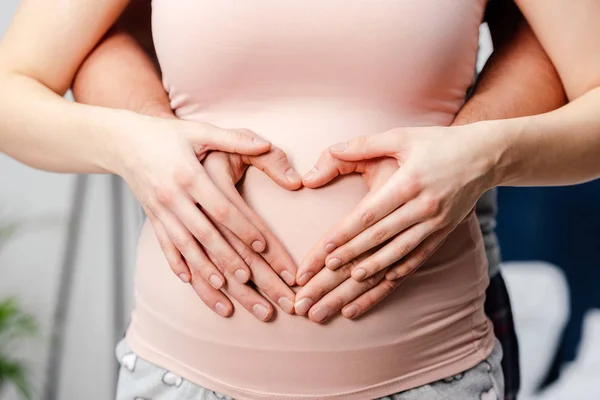 Cropped shot of young pregnant couple touching belly and showing hand heart symbol — Stock Photo