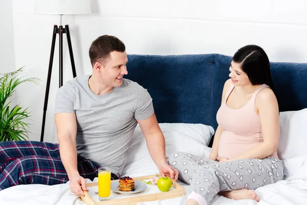 Giovane uomo sorridente che tiene vassoio con la prima colazione e guardando felice donna incinta a letto — Foto stock