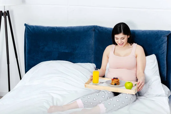 Smiling young pregnant woman holding tray with breakfast in bed — Stock Photo