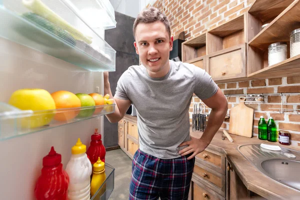 Guapo joven sonriente en pijama mirando en el refrigerador - foto de stock