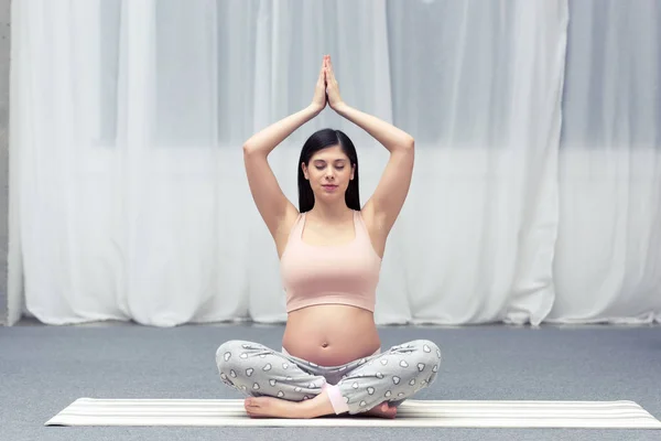 Joven embarazada sentada en la esterilla y practicando yoga en casa - foto de stock