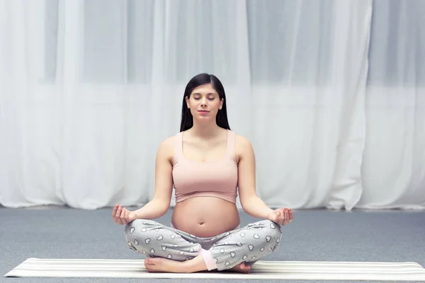 Jeune femme enceinte avec les yeux fermés assis en position lotus sur tapis de yoga — Photo de stock