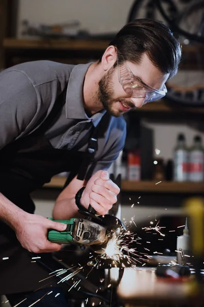 Joven con gafas protectoras cortando metal con amoladora angular en taller - foto de stock