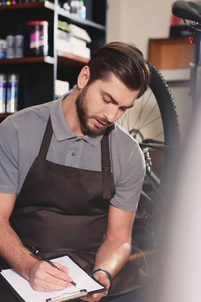 Focused young worker in apron taking notes on clipboard at bicycle shop — Stock Photo