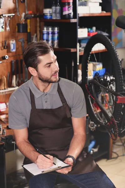 Красивый молодой велосипедный механик сидит и делает заметки в мастерской — стоковое фото