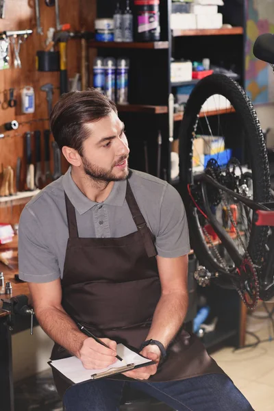 Bello giovane meccanico di biciclette seduto e prendere appunti in officina — Foto stock