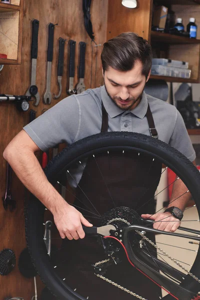 Young man in apron fixing bicycle wheel and chain with wrench in workshop — Stock Photo
