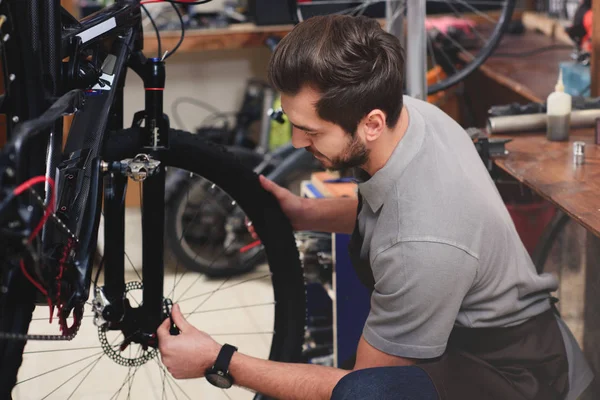 Bonito jovem mecânico no avental fixação roda de bicicleta na oficina — Fotografia de Stock