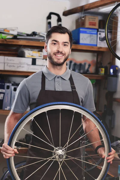 Joven trabajador guapo en delantal sosteniendo rueda de bicicleta y sonriendo a la cámara en el taller - foto de stock