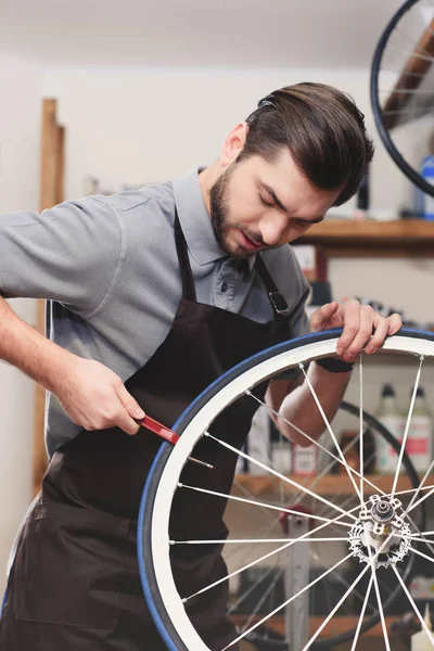 Beau jeune travailleur dans tablier de fixation roue de vélo dans l'atelier — Photo de stock