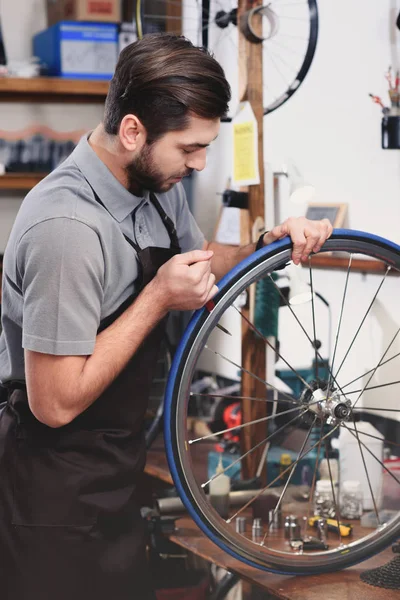 Joven mecánico en delantal reparando rueda de bicicleta en taller - foto de stock