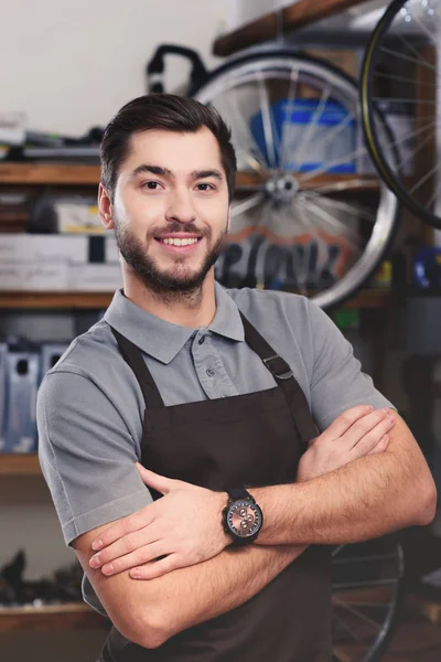 Handsome young bicycle mechanic in apron standing with crossed arms and smiling at camera — Stock Photo