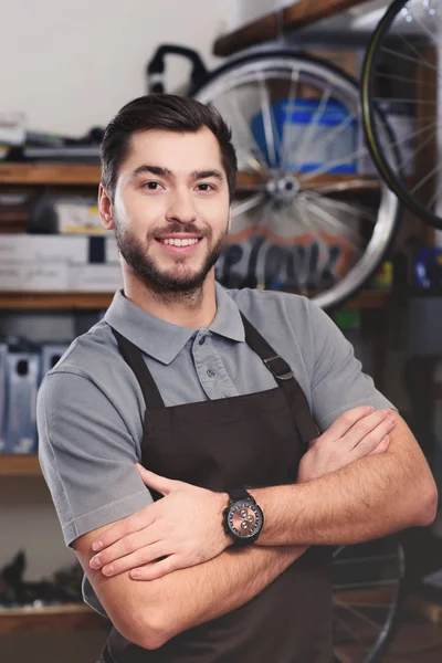 Bel giovane meccanico di biciclette in grembiule in piedi con le braccia incrociate e sorridente alla macchina fotografica — Foto stock